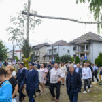 Grupa ludzi w formalnych strojach idzie trawnikiem. W tle są domy oraz duży, poziomy pień na słupie. Otoczenie jest zielone i słoneczne.