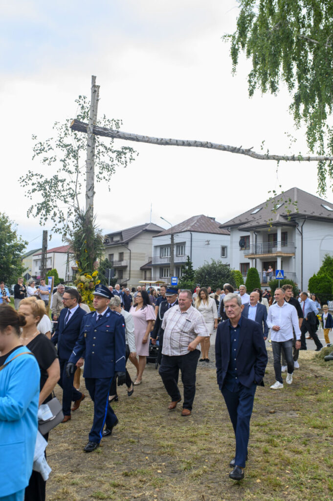 Grupa ludzi w formalnych strojach idzie trawnikiem. W tle są domy oraz duży, poziomy pień na słupie. Otoczenie jest zielone i słoneczne.