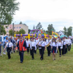 Orkiestra maszeruje po zielonej trawie podczas festynu. Członkowie ubrani w biało-czarne stroje. W tle kolorowe dmuchane zamki.