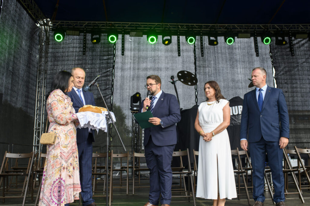 Na scenie stoi pięć osób. Kobieta trzyma chleb na białej serwecie. Tło ciemne, oświetlenie zielonymi reflektorami.