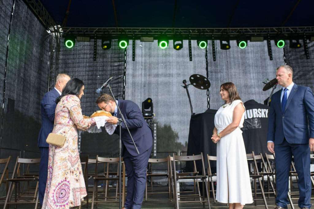Na scenie trwa uroczystość; mężczyzna w garniturze składa pocałunek na bochenku chleba. Kobieta w kolorowej sukience go podaje. Tło to ciemna kurtyna i zielone światła sceniczne.