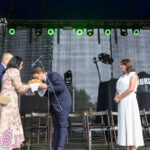 Na scenie trwa uroczystość; mężczyzna w garniturze składa pocałunek na bochenku chleba. Kobieta w kolorowej sukience go podaje. Tło to ciemna kurtyna i zielone światła sceniczne.