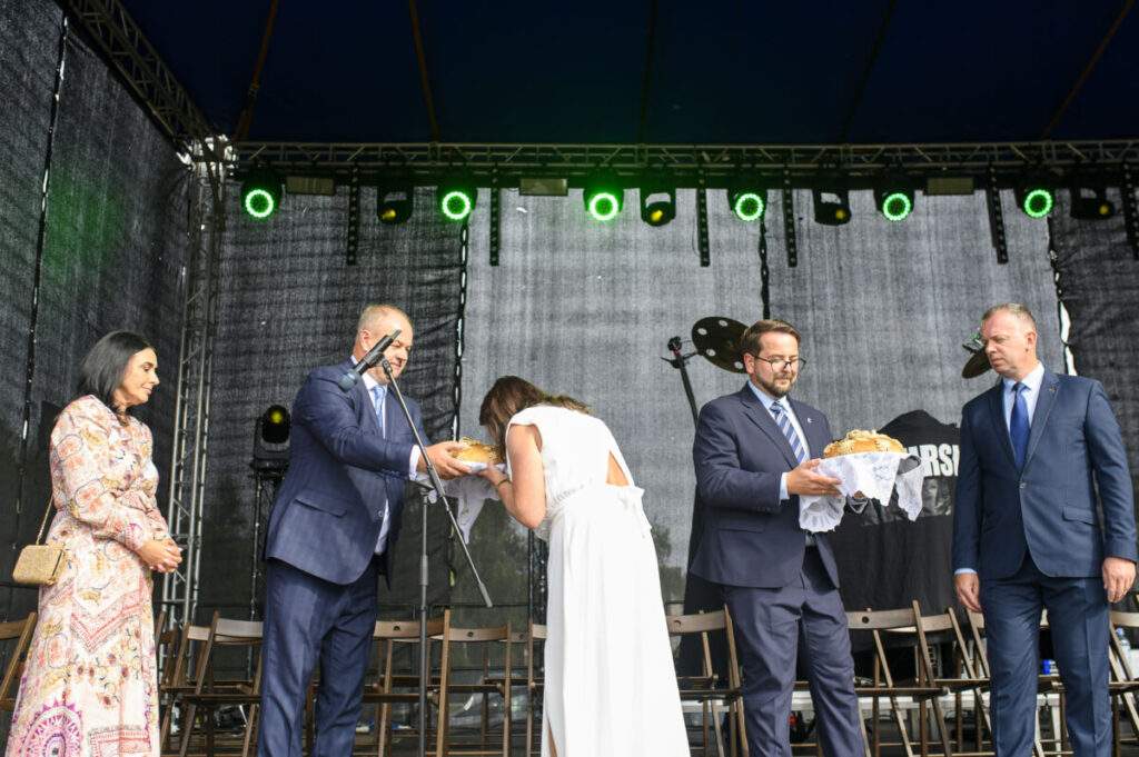 Grupa ludzi na scenie przy chlebach, elegancko ubrani, w tle czarny materiał i zielone światła sceny.