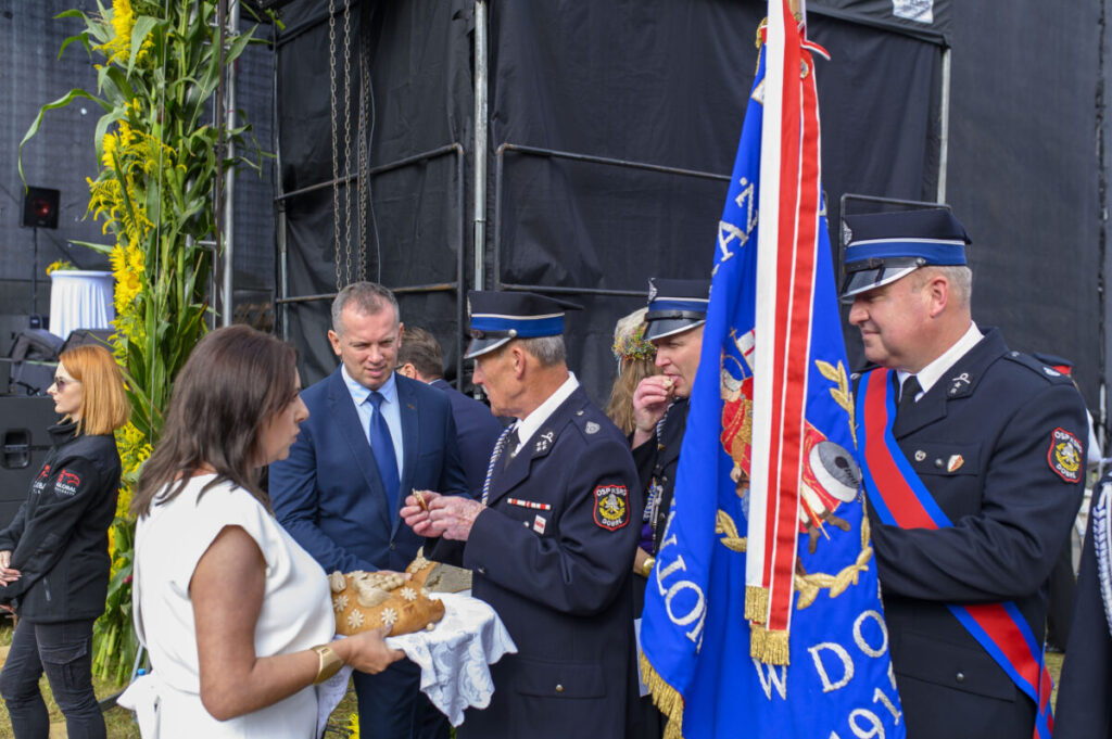 Na zdjęciu widać osoby w uroczystych mundurach i narodową flagę. Po lewej kobieta trzyma tradycyjny chleb. Tło stanowią wysokie słoneczniki i czarne dekoracje.