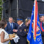 Na zdjęciu widać osoby w uroczystych mundurach i narodową flagę. Po lewej kobieta trzyma tradycyjny chleb. Tło stanowią wysokie słoneczniki i czarne dekoracje.