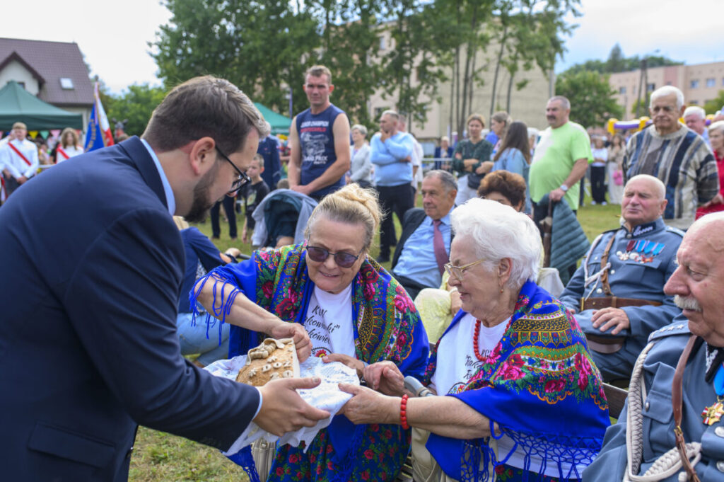 Na zdjęciu starsze kobiety w kolorowych chustach przekazują chleb mężczyźnie. W tle zielone drzewa i ludzie. Uroczystość na świeżym powietrzu.