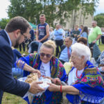 Na zdjęciu starsze kobiety w kolorowych chustach przekazują chleb mężczyźnie. W tle zielone drzewa i ludzie. Uroczystość na świeżym powietrzu.