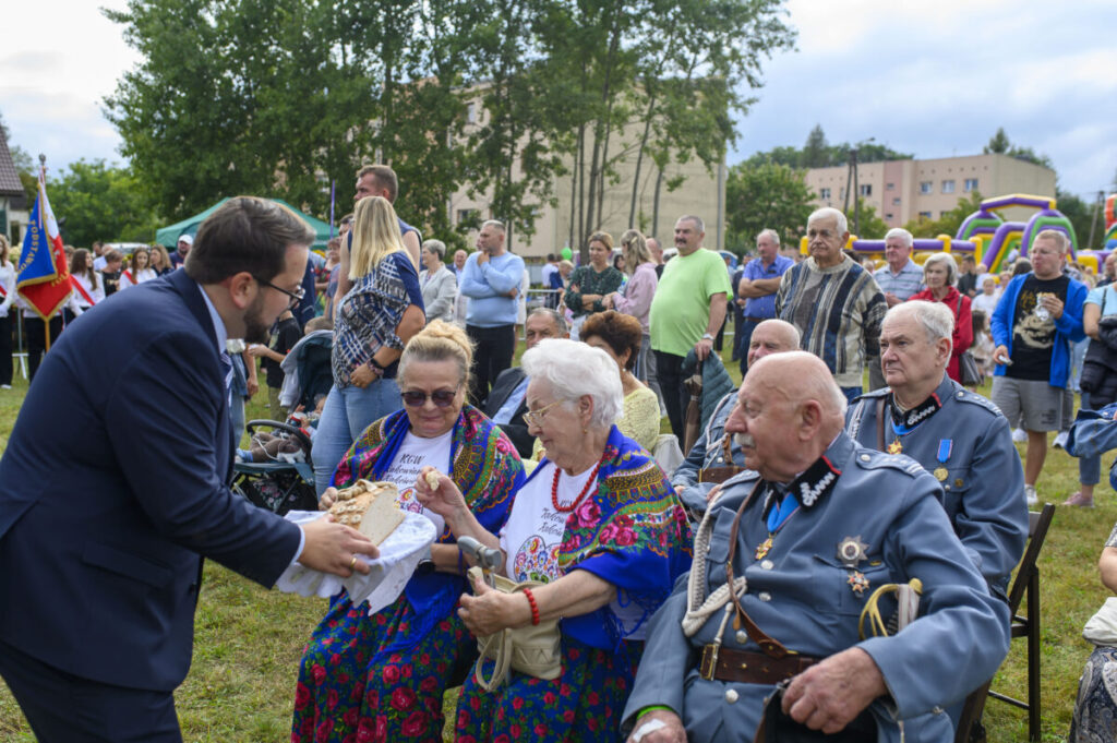 Na zdjęciu grupa ludzi na zewnątrz, mężczyzna w garniturze wręcza chleb kobiecie w kolorowym stroju ludowym. W tle tłum ludzi, namioty i drzewa. Dzień jest pochmurny.