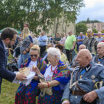 Na zdjęciu grupa ludzi na zewnątrz, mężczyzna w garniturze wręcza chleb kobiecie w kolorowym stroju ludowym. W tle tłum ludzi, namioty i drzewa. Dzień jest pochmurny.