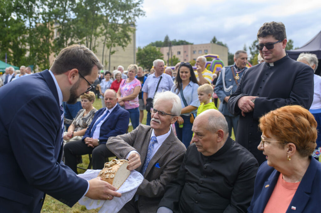 Grupa ludzi siedzi na trawie podczas wydarzenia plenerowego. Mężczyzna z brodą w garniturze podaje chleb mężczyźnie w okularach. W tle widać drzewa i tłum osób w różnorodnych strojach.