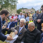 Grupa ludzi siedzi na trawie podczas wydarzenia plenerowego. Mężczyzna z brodą w garniturze podaje chleb mężczyźnie w okularach. W tle widać drzewa i tłum osób w różnorodnych strojach.