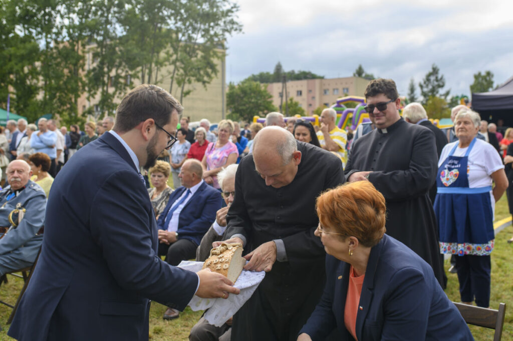 Na zdjęciu widoczni są ludzie na zewnątrz podczas wydarzenia. Starszy duchowny trzyma chleb, obok niego inne osoby. W tle widać zielone drzewa i budynki.