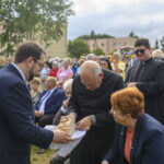 Na zdjęciu widoczni są ludzie na zewnątrz podczas wydarzenia. Starszy duchowny trzyma chleb, obok niego inne osoby. W tle widać zielone drzewa i budynki.