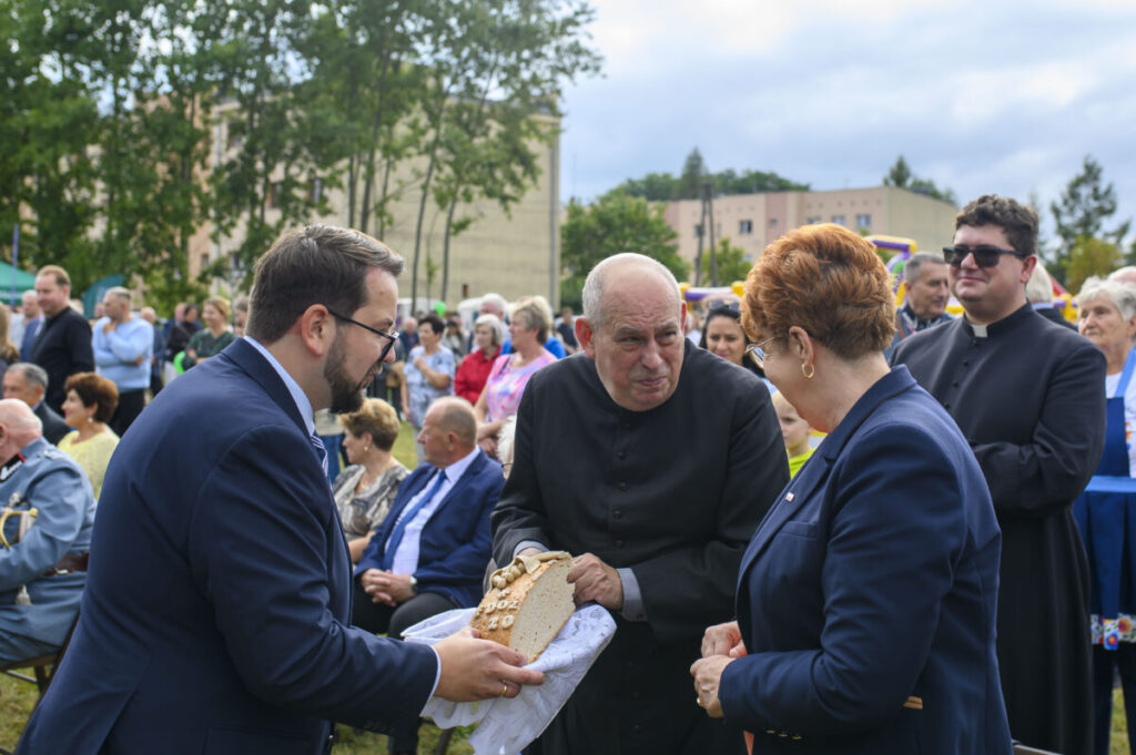 Na zdjęciu grupa osób na wydarzeniu plenerowym. Mężczyzna w czerni trzyma chleb. W tle drzewa i ludzie w kolorowych ubraniach.