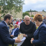 Na zdjęciu grupa osób na wydarzeniu plenerowym. Mężczyzna w czerni trzyma chleb. W tle drzewa i ludzie w kolorowych ubraniach.
