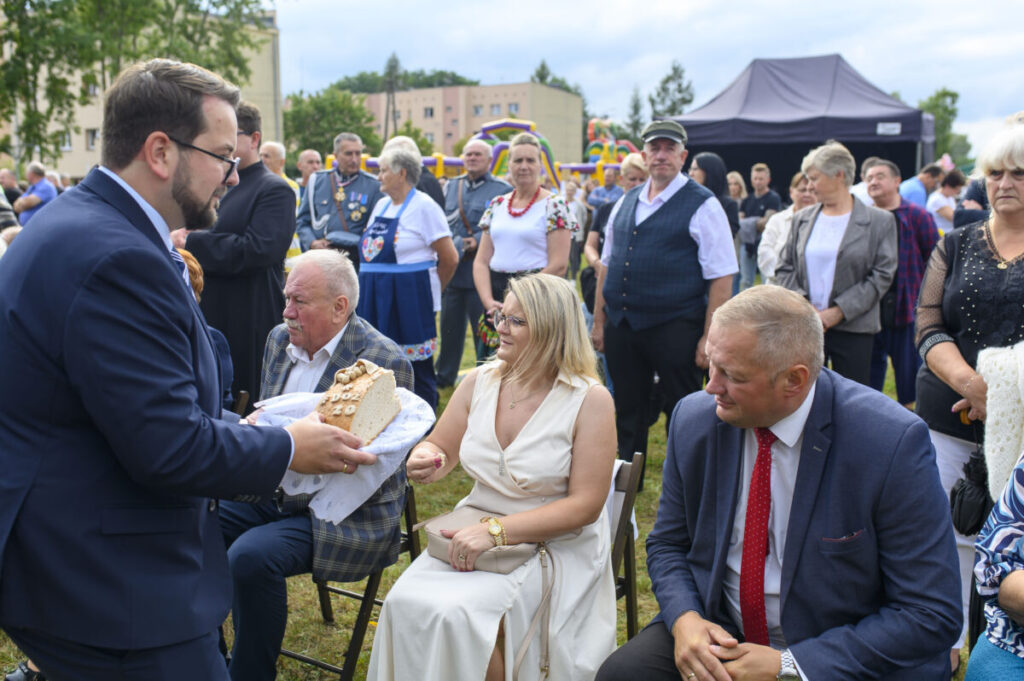 Na zdjęciu mężczyzna w garniturze oferuje chleb osobom siedzącym na krzesłach. Tło zielone, na zewnątrz, z namiotem i tłumem. Ludzie ubrani elegancko, atmosfera formalna.