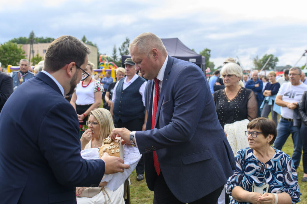 Na zdjęciu grupa osób w plenerze; mężczyzna podaje chleb drugiemu. W tle widać niebo, namiot i kolorowe ubrania. Atmosfera przypomina wydarzenie na świeżym powietrzu.