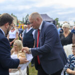 Na zdjęciu grupa osób w plenerze; mężczyzna podaje chleb drugiemu. W tle widać niebo, namiot i kolorowe ubrania. Atmosfera przypomina wydarzenie na świeżym powietrzu.