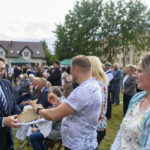 Na obrazie widoczna jest grupa ludzi na zewnątrz. Mężczyzna w garniturze trzyma chleb, który inna osoba kroi. W tle widać domy i drzewa.