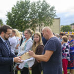 Grupa ludzi stoi na zewnątrz; mężczyzna w garniturze podaje chleb. W tle drzewa i namiot. Atmosfera festynu.