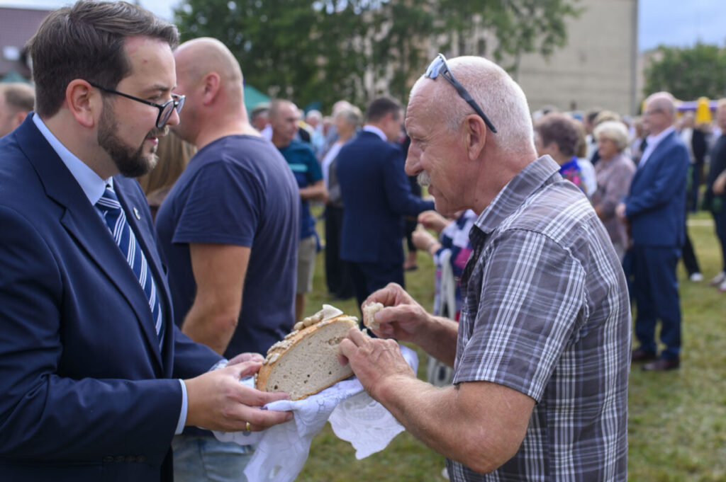 Mężczyzna w garniturze podaje chleb starszemu mężczyźnie w kratkowanej koszuli na zewnątrz. W tle ludzie i drzewa. Trawiaste podłoże.