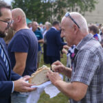 Mężczyzna w garniturze podaje chleb starszemu mężczyźnie w kratkowanej koszuli na zewnątrz. W tle ludzie i drzewa. Trawiaste podłoże.