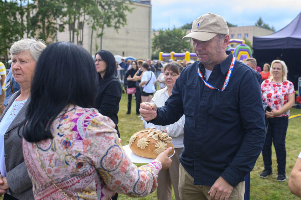 Na zdjęciu ludzie na zewnątrz, w tym mężczyzna z chlebem ozdobionym kwiatami. Tło to drzewa i namioty. Kolory są żywe, dominują zielenie i pastele.