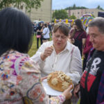 Na zdjęciu ludzie na zewnątrz, trzymają chleb ozdobiony kwiatami. Kobieta w białej bluzce sięga po kromkę. W tle zielone drzewa i ludzi.