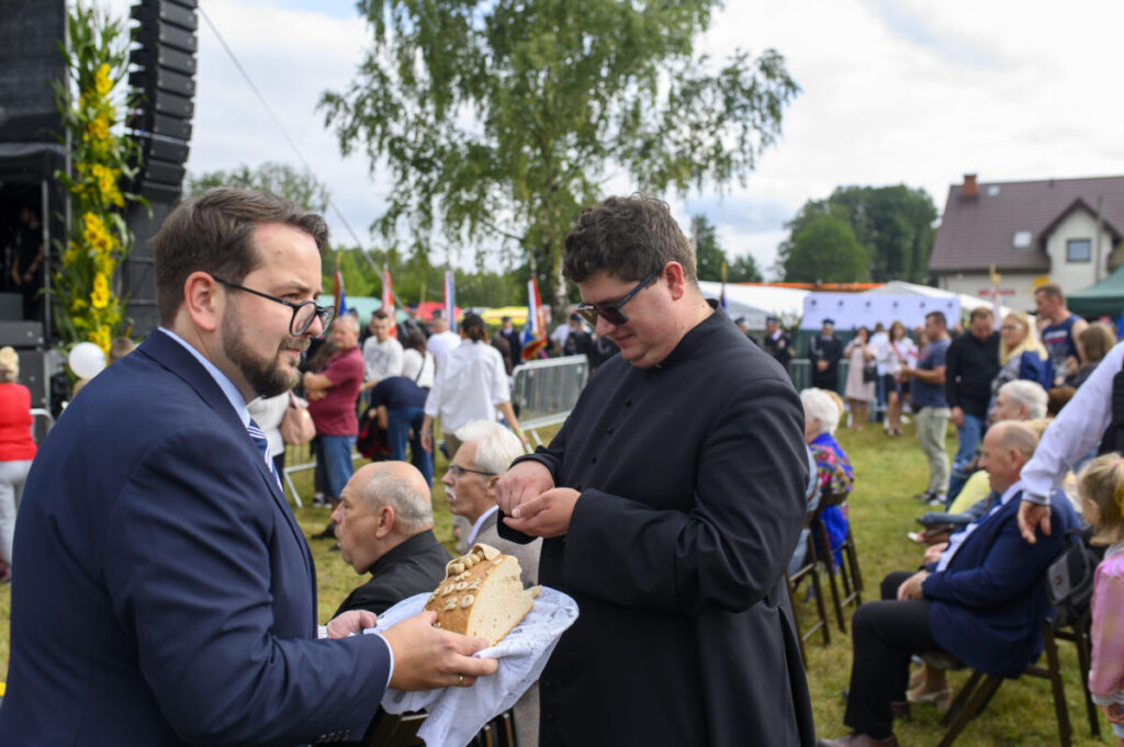 Na zdjęciu dwóch mężczyzn, jeden w garniturze podejmuje chleb, drugi w ciemnym stroju. W tle tłum ludzi i zielone drzewa. Atmosfera festiwalowa, jasne kolory.