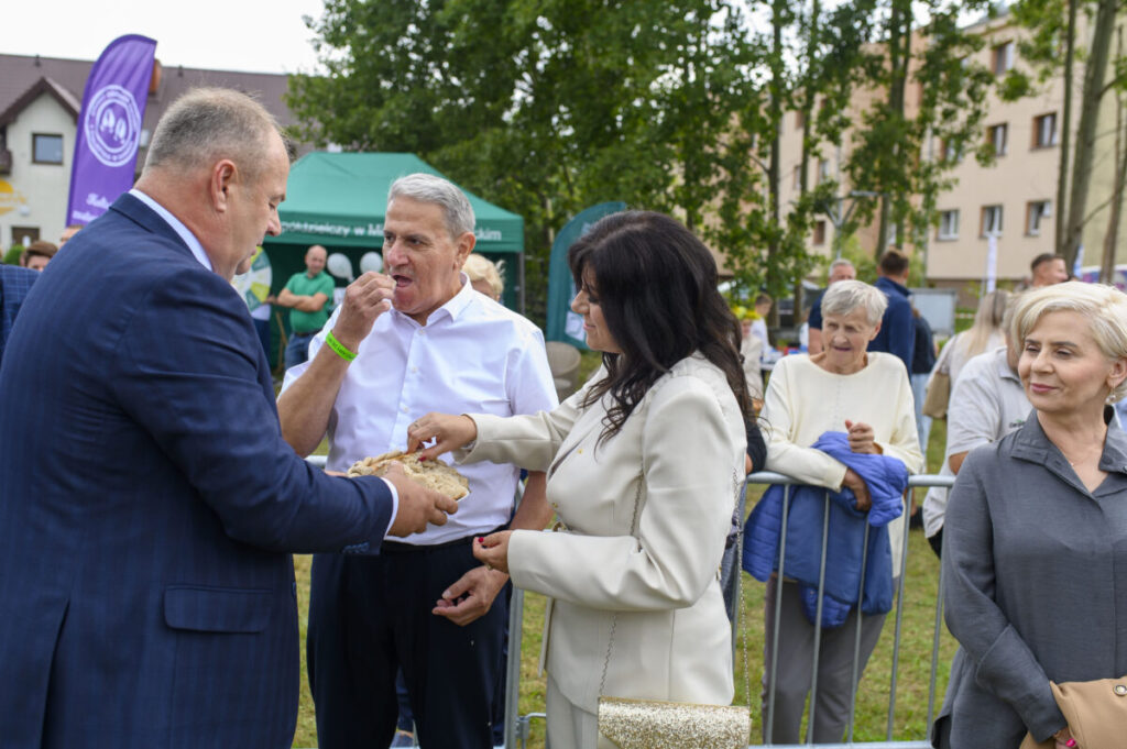 Grupka dzieci idzie w parach machają ręką dwie panie. W tle biały budynek i drewniany ganek.