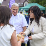 Na zdjęciu grupa ludzi, kobieta wręcza chleb mężczyźnie w białej koszuli. Tło zdobią zielone namioty i drzewa. Atmosfera pikniku na świeżym powietrzu.