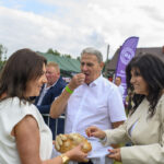 Na zdjęciu widać ludzi na zewnątrz w jasnych ubraniach, dzielących się chlebem. W tle zielone namioty i fioletowe banery. Krajobraz jest pogodny, z drzewami i niebieskim niebem.
