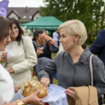 Na zdjęciu grupa ludzi podczas wydarzenia na świeżym powietrzu. Kobieta podaje chleb; w tle zielony namiot i drzewa. Panuje pogodna atmosfera.