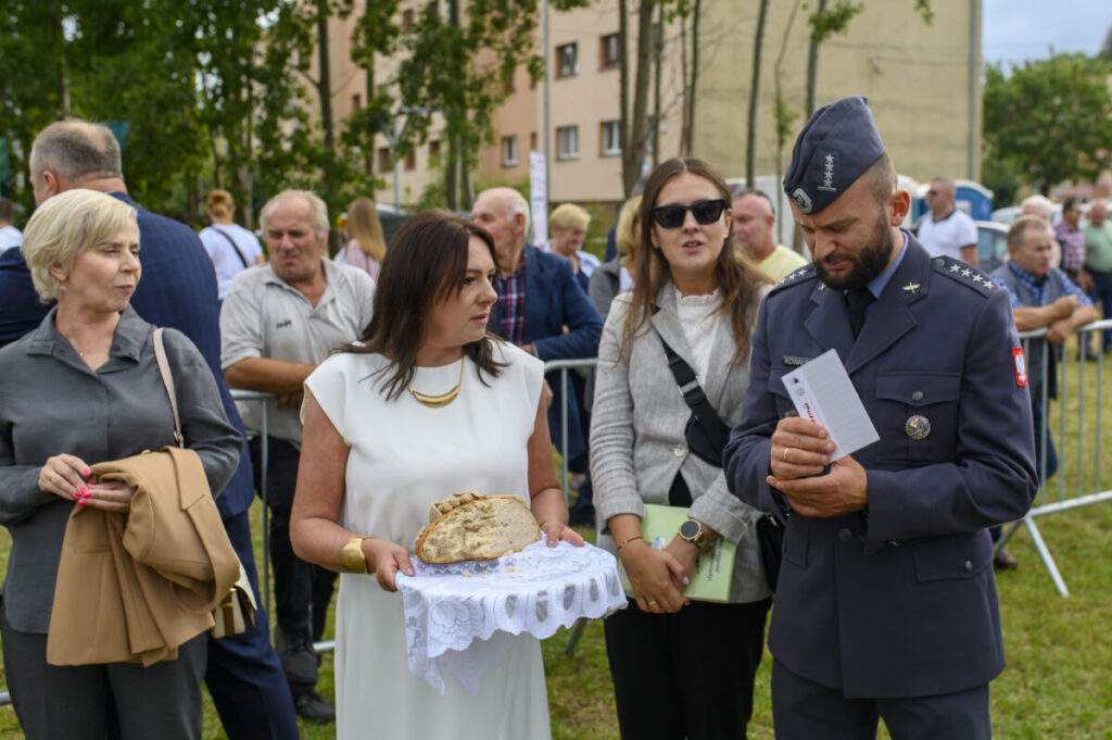 Grupa ludzi uczestniczy w wydarzeniu na zewnątrz. Kobieta w białej sukni trzyma chleb na ozdobnej serwetce. W tle są drzewa i budynki.