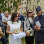 Grupa ludzi uczestniczy w wydarzeniu na zewnątrz. Kobieta w białej sukni trzyma chleb na ozdobnej serwetce. W tle są drzewa i budynki.