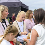 Grupa osób na świeżym powietrzu przed czarnym namiotem. Kobiety i dzieci trzymają koszyk z chlebem. W tle zielona trawa i niebo.