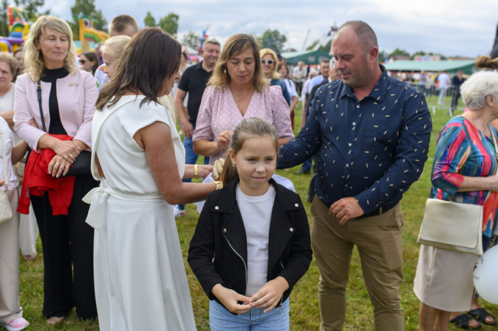 Grupa ludzi na zewnątrz, zielona trawa i kolorowe dmuchane zamki. Dziewczynka w czarnej kurtce na pierwszym planie. W tle tłum i namioty.