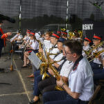 Orkiestra dęta w granatowych strojach gra na scenie. Muzycy trzymają instrumenty dęte, jak saksofony i trąbki. Tło ciemne, widoczny dyrygent z przodu.