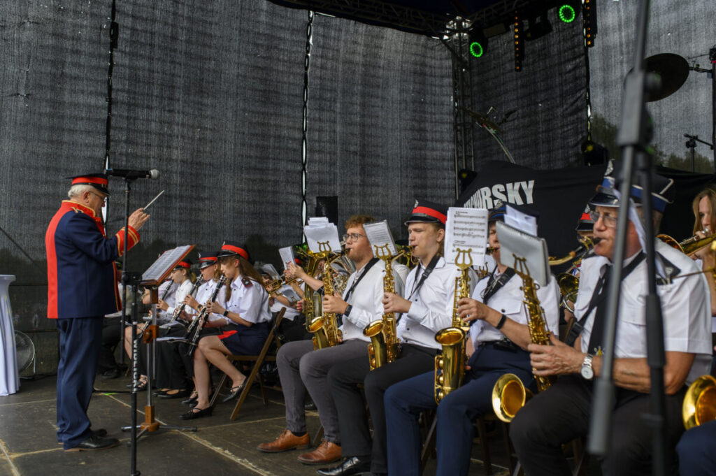 Orkiestra dęta na scenie, muzycy w białych koszulach i czerwonych czapkach. Dyrygent w granatowo-czerwonym mundurze. Czarne tło.