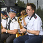 Orkiestra dęta z muzykami w białych mundurach gra na scenie. Tło to ciemna kurtyna i drzewa. Instrumenty są złote.