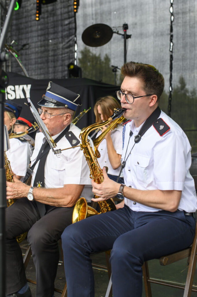 Orkiestra dęta z muzykami w białych mundurach gra na scenie. Tło to ciemna kurtyna i drzewa. Instrumenty są złote.