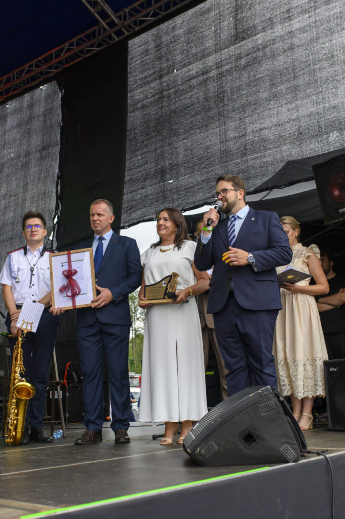 Grupa osób na scenie, w eleganckich ubraniach, trzyma nagrody. Tło to ciemny ekran. Obok stoi mężczyzna z mikrofonem.