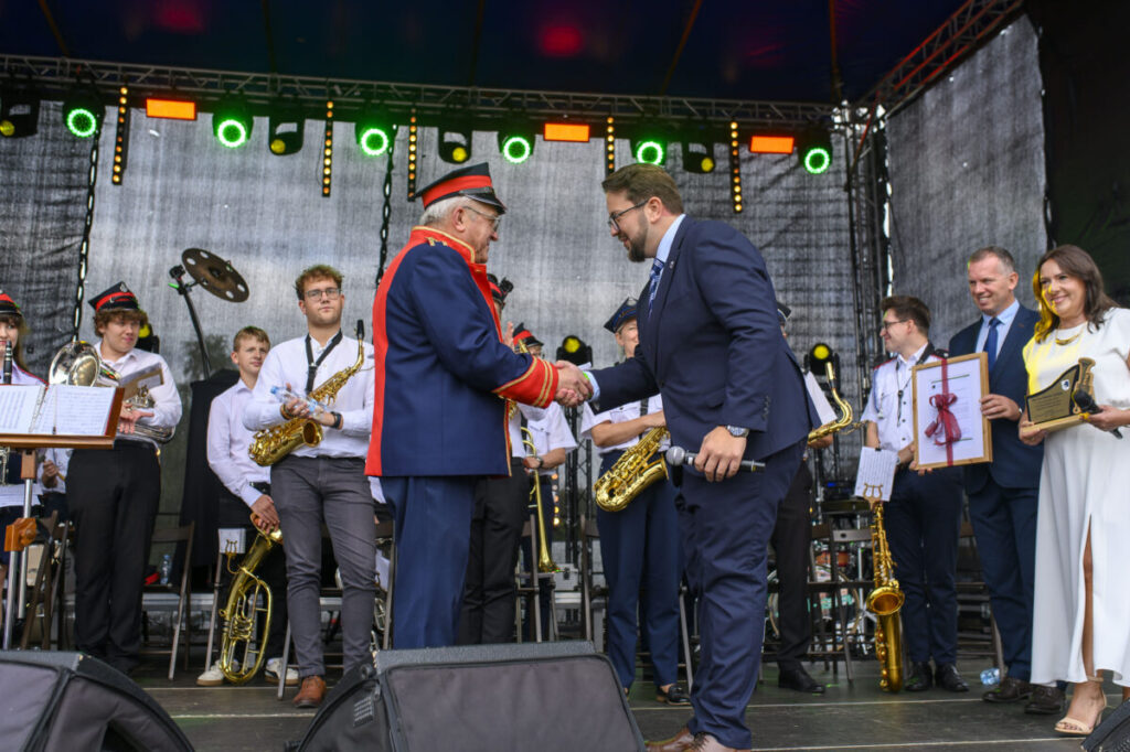 Na scenie orkiestra, mężczyźni w mundurach podają sobie ręce. Światła zielone i pomarańczowe w tle. Kobieta trzyma dyplom.