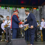 Na scenie orkiestra, mężczyźni w mundurach podają sobie ręce. Światła zielone i pomarańczowe w tle. Kobieta trzyma dyplom.