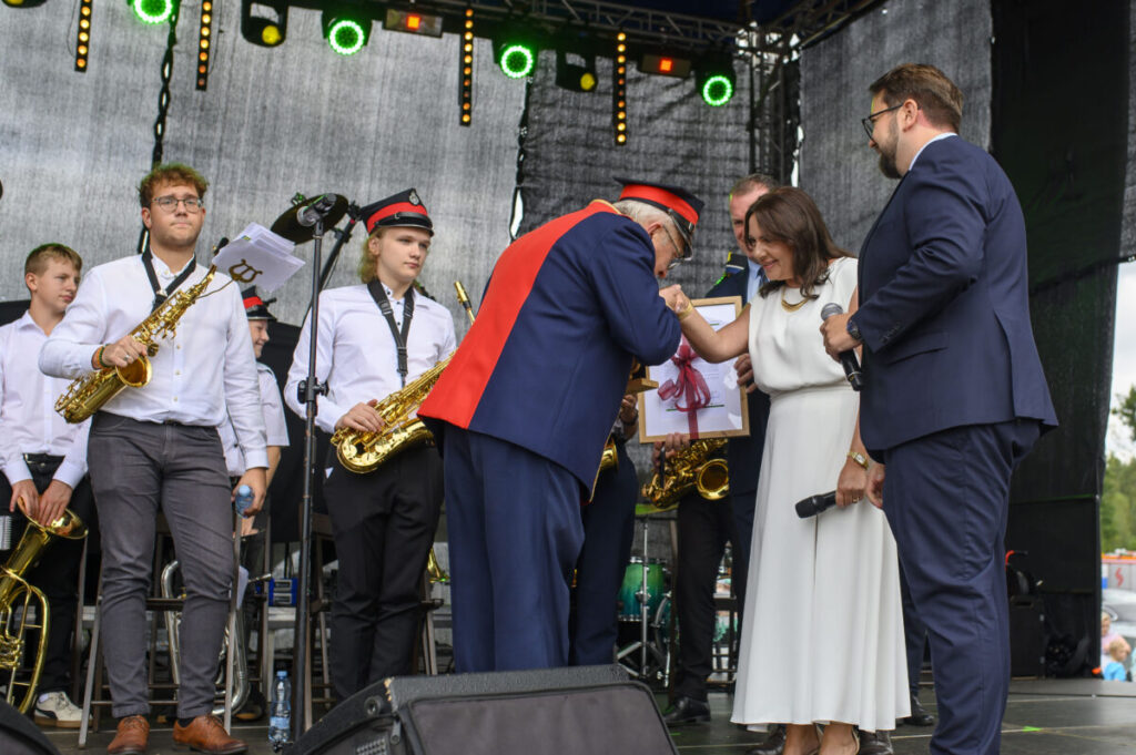 Na scenie stoi orkiestra dęta w białych koszulach i ciemnych spodniach. Mężczyzna w stroju z czerwonymi akcentami wita się z kobietą w białej sukni. Tło jest ciemne, z kolorowymi światłami.