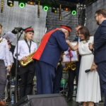 Na scenie stoi orkiestra dęta w białych koszulach i ciemnych spodniach. Mężczyzna w stroju z czerwonymi akcentami wita się z kobietą w białej sukni. Tło jest ciemne, z kolorowymi światłami.