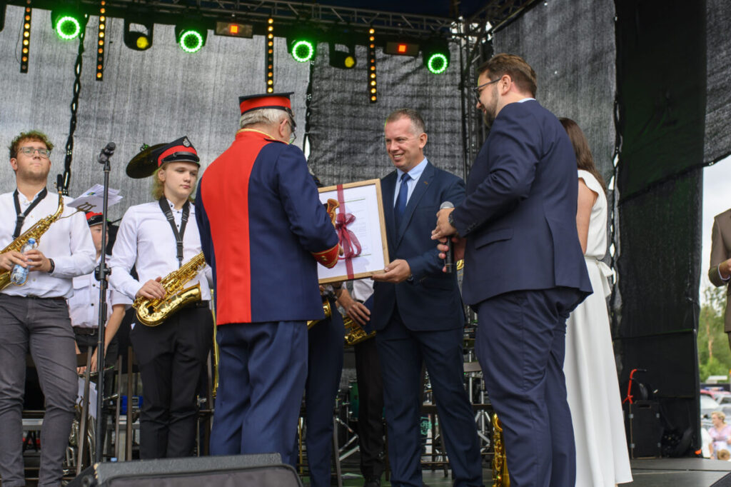 Na scenie mężczyzna w mundurze wręcza dyplom innemu mężczyźnie w garniturze. Obok stoją muzycy z saksofonami, ubrani na biało i czarno. Tło to ciemne zasłony i kolorowe światła sceniczne.
