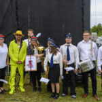 Grupa osób z instrumentami muzycznymi stoi na trawie. Jedna osoba w jaskrawozielonym garniturze. Tło jest czarne.