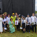 Grupa ludzi na tle sceny na świeżym powietrzu. Ubrani w różnorodne stroje, trzymają instrumenty muzyczne. Tło jest ciemne, trawiasta ziemia.