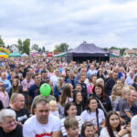 Tłum ludzi na festynie. Kolorowe dmuchańce w tle. Pogoda pochmurna, ludzie patrzą w stronę sceny.